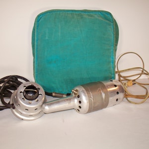 May include: A vintage handheld electric massager with a metallic silver cylindrical body featuring ventilation holes, a black power cord, and a separate circular attachment. A square, teal-colored fabric cushion is positioned behind the device.