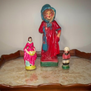 May include: Three vintage figurines on a wooden tray. A girl in a red dress and blue hooded cloak, a girl in a pink dress, and a boy in a blue and white striped shirt and red pants.