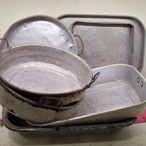 May include: A collection of vintage metal cookware, including a large pot with handles, a round lid, and several rectangular baking pans. The pieces have a worn, silver finish, suggesting age and use. The arrangement is stacked, showcasing the variety of shapes and sizes.