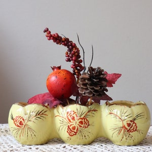 May include: A yellow ceramic planter with a three-section design features a gold and red pine cone pattern. The planter is filled with a small arrangement of red berries, a red pomegranate, and a brown pine cone.