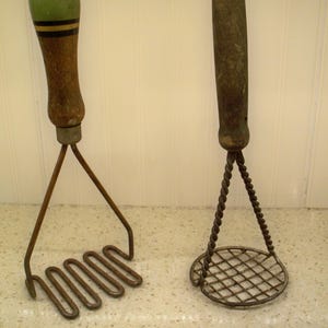 May include: Two vintage potato mashers. One has a green handle with black and yellow stripes and a wavy metal head. The other has a twisted metal handle and a circular wire grid head. Both are made of wood and metal.