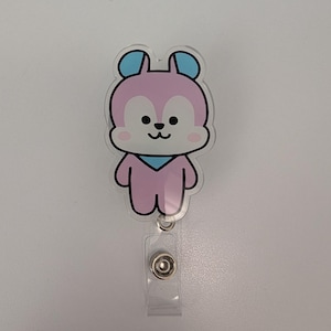 May include: A pink and blue cartoon bear badge reel. The bear has a black outline, blue ears, and a blue scarf. The badge reel has a clear plastic strap and a metal snap.