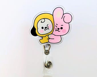 May include: An acrylic badge reel featuring a cartoon illustration of a yellow character in a hood hugging a pink rabbit character. The design is attached to a clear plastic strap with a metal snap button and a metal swivel hook for attachment.