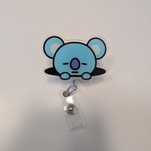May include: A light blue koala bear design on a retractable badge reel. The koala has large ears and a sleepy expression. The reel has a clear plastic strap and a metal clip for attaching to clothing or a bag.
