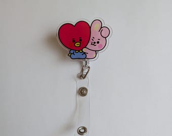 May include: A retractable badge reel featuring a clear acrylic topper with a graphic of a red heart-shaped character wearing a blue outfit hugging a light pink rabbit character. The reel includes a clear plastic strap with two silver metal snaps.