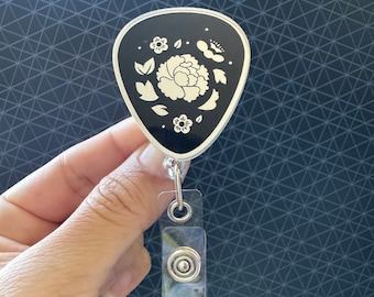 May include: A black badge reel with a guitar pick shape featuring a white floral design. The badge reel includes a clear plastic strap with a silver metal snap button closure, designed to hold an identification card or badge securely in place.