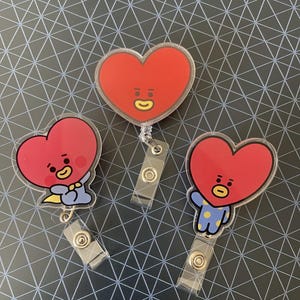 May include: Three red heart-shaped badge reels with cartoon faces. The badge reels have clear plastic bodies and metal clips. One heart has a pacifier, one is waving, and one has a smiling face.
