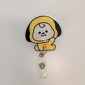 May include: A yellow and black cartoon character badge reel with a clear plastic strap and a metal clip. The character is wearing a yellow hooded outfit with red buttons and has a smiling face. The badge reel is on a white background.
