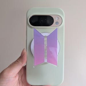 Könnte beinhalten: Eine mintgrüne Handyhülle mit einem Farbverlauf-Logo in Lila und Rosa. Das Logo ist ein stilisiertes Design. Die Handyhülle hat eine Kamera und einen Blitz. Das Telefon wird von einer Hand gehalten.