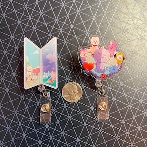 May include: Two clear acrylic badge reels with cartoon character designs. One reel features an open book design with a sky and character scene. The other reel has a group of cartoon characters. A quarter is next to the reels.
