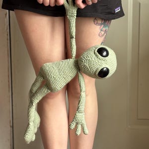 Op de afbeelding: Een gehaakte alienpop in lichtgroen, met grote zwarte ogen en langwerpige ledematen. Het handgemaakte pluche speelgoed wordt bij de arm vastgehouden. De alien heeft een afgeronde kop en gedetailleerde handen en voeten.