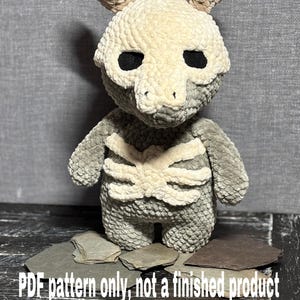 Könnte beinhalten: Ein gehäkeltes Plüschtier in Form eines Skeletts mit Geweih. Das Spielzeug ist grau und weiß mit schwarzen Augen. Das Spielzeug sitzt auf einem Stapel Schieferplatten. Der Text "PDF pattern only, not a finished product" befindet sich am unteren Rand des Bildes.