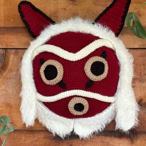 Op de afbeelding: Een gehaakt pluche masker in de vorm van een rood en wit geestmasker met zwarte ogen en een witte bontkraag.
