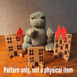May include: A crochet pattern for a gray Godzilla monster with red flames on the sides of the cardboard buildings. The pattern is for a toy and not a physical item.