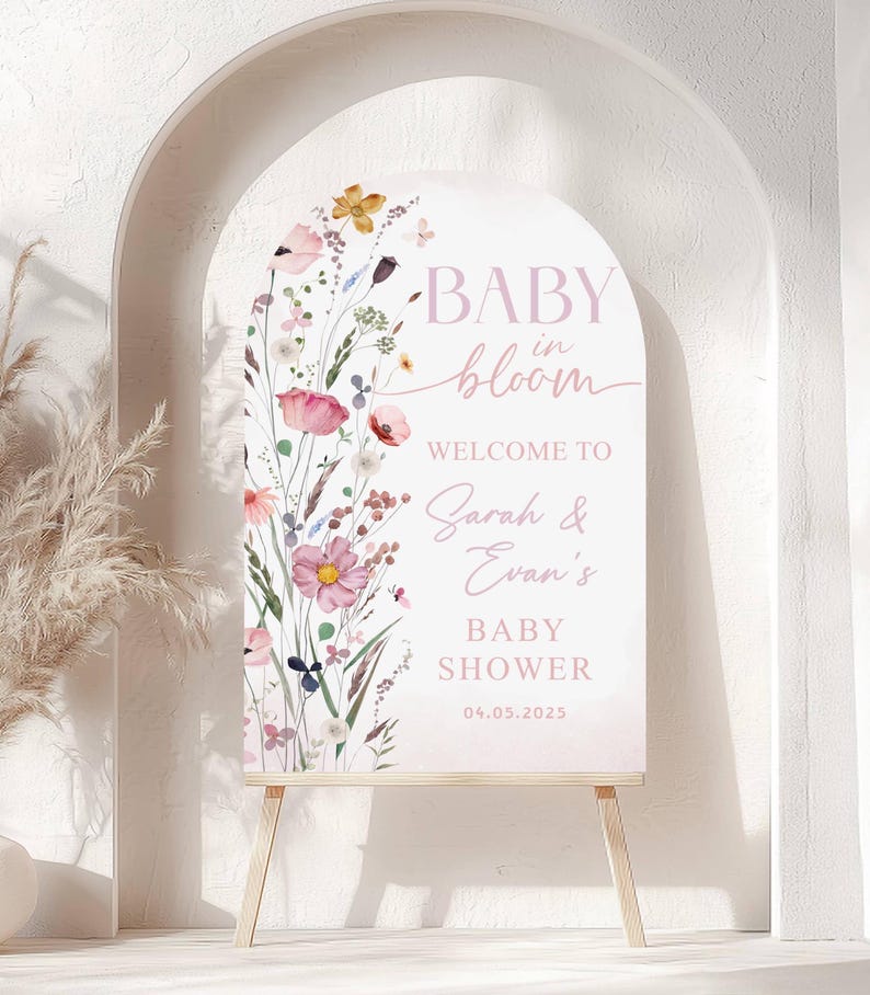 May include: A baby shower welcome sign with a floral design. The sign reads "BABY in bloom" and welcomes guests to Sarah & Evan's baby shower on 04.05.2025. The sign is displayed on a wooden easel.