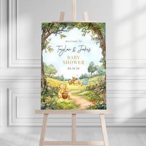 May include: Welcome to Taylor & Jake's Baby Shower sign with a Winnie the Pooh theme. The sign features a watercolor illustration of Winnie the Pooh, Piglet, and Eeyore in a forest setting. The text "Welcome to Taylor & Jake's Baby Shower 06.16.24" is written in gold lettering.