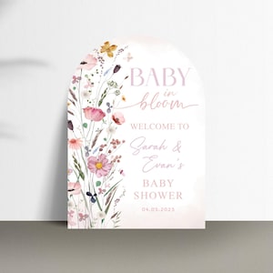 May include: A baby shower sign with a floral design. The sign is arched at the top and features the words "BABY in bloom" and "WELCOME TO Sarah & Evan's BABY SHOWER 04.03.2025". The sign is decorated with pink, yellow, and purple flowers.