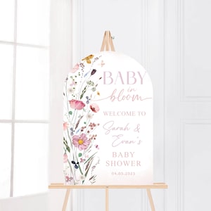May include: A white, arched baby shower welcome sign with floral watercolor illustrations. The sign reads "BABY in bloom" and welcomes guests to Sarah & Evan's baby shower on 04.05.2025. The sign is displayed on a wooden easel.