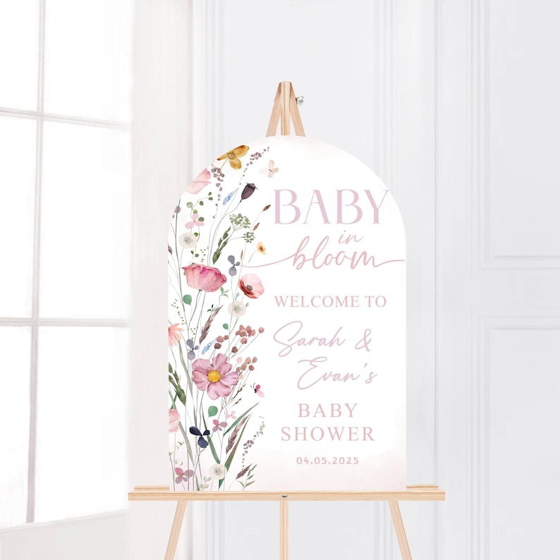 May include: A white, arched baby shower welcome sign with floral watercolor illustrations. The sign reads "BABY in bloom" and welcomes guests to Sarah & Evan's baby shower on 04.05.2025. The sign is displayed on a wooden easel.