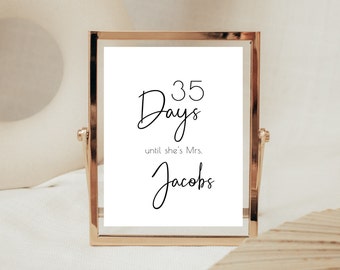 May include: A framed sign with gold trim displays the text "35 Days until she's Mrs. Jacobs" in elegant black script. The sign is set against a soft, neutral background, suggesting a wedding countdown theme.