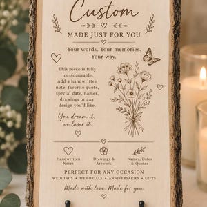Puede incluir: Una placa rectangular de madera clara con bordes de corteza natural, exhibida sobre un soporte de metal negro. La superficie presenta texto e ilustraciones florales grabados con láser, incluyendo una mariposa y un ramo de flores silvestres.