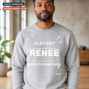 Può includere: Una felpa girocollo grigio melange con testo bianco che recita CLAY DAY, RENEE e MADE BY HAND 2026. Il design include semplici linee curve e trattini bianchi che circondano il testo, creando un'estetica grafica pulita ed essenziale.