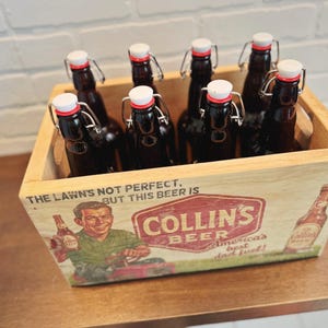 May include: A wooden crate filled with eight dark glass beer bottles with white and red stoppers. The crate features a vintage Collins Beer advertisement with the text "The Lawn's Not Perfect, But This Beer Is."