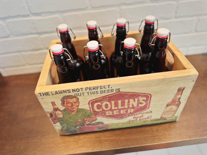 May include: A wooden crate filled with eight dark glass beer bottles with white and red stoppers. The crate features a vintage Collins Beer advertisement with the text "The Lawn's Not Perfect, But This Beer Is."
