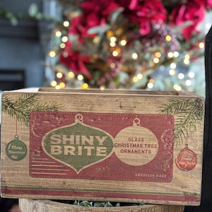 Könnte beinhalten: Eine Holzkiste mit einem roten, grünen und weißen Etikett, auf dem "Shiny Brite Glass Christmas Tree Ornaments American Made" steht. Die Kiste steht auf einer Metalloberfläche.