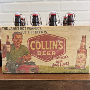 May include: Wooden beer carrier with six brown glass bottles. The carrier features a vintage-style graphic with the text "COLLIN'S BEER" and "America's best dad fuel!" The bottles have white and red stoppers.