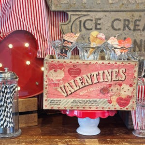 May include: A vintage wooden box filled with Valentine's Day cards. The box is decorated with red hearts and the words "Valentines with Envelopes". The box is sitting on a wooden table with a red and white striped tablecloth in the background.
