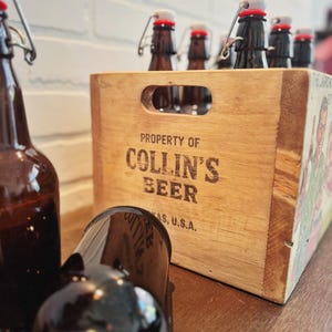 May include: A wooden crate labeled "PROPERTY OF COLLIN'S BEER, TEXAS, U.S.A." holds several brown glass bottles with swing-top closures. One bottle lies in front of the crate. The crate is on a wooden surface with a white brick wall in the background.