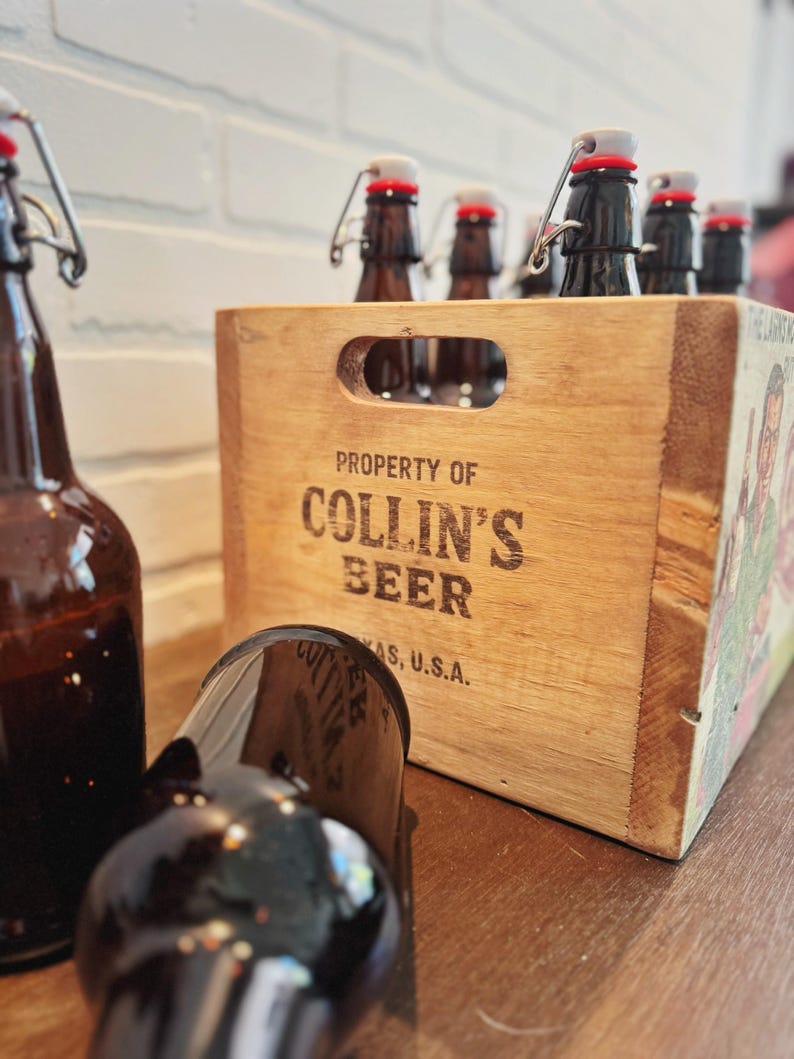 May include: A wooden crate labeled "PROPERTY OF COLLIN'S BEER, TEXAS, U.S.A." holds several brown glass bottles with swing-top closures. One bottle lies in front of the crate. The crate is on a wooden surface with a white brick wall in the background.