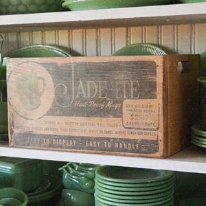 May include: A collection of jadeite green dishware, including plates, bowls, and mugs, displayed on shelves. A wooden crate with the text "JADEITE Heat Proof Mugs" is visible. The dishes are stacked and arranged, showcasing a vintage aesthetic.