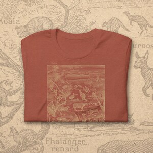 Può includere: T-shirt color ruggine con un'illustrazione dettagliata della fauna australiana nei toni del rosso e beige. Il design include canguri, emù e altri animali. La maglietta ha un girocollo classico e maniche corte.