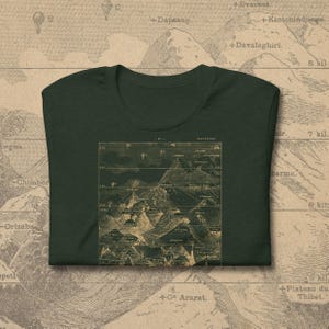 May include: Dark green t-shirt featuring a vintage map design in tan and brown tones. The map depicts mountain ranges with labels and coordinates. The shirt is made of soft fabric and has a classic crew neck.