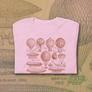 May include: A pink t-shirt featuring a vintage illustration of various hot air balloons and airships. The illustration is in brown ink and includes text in French. The text reads "Ballon sphérique (1918)".