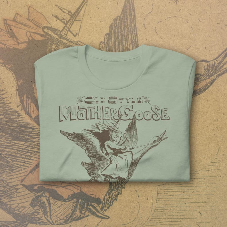 May include: A light green t-shirt with the words "OLD STYLE MOTHER GOOSE" in brown lettering. The shirt features a brown illustration of a woman riding a goose. The shirt is folded.