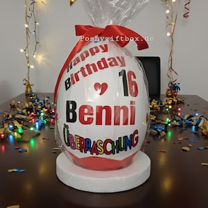 Może przedstawiać: Biały, owalny pakunek prezentowy z napisem "Happy Birthday 16 Benni &Uuml;berraschung" w kolorze czerwonym i czarnym. Pakunek stoi na białym stojaku, otoczony konfetti i balonami w kolorze złotym, niebieskim i czerwonym.