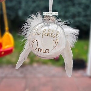 May include: A clear glass ornament with white feathers attached. The ornament has the text "Du fehlst Oma" written in gold lettering with a gold heart.