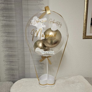 May include: A clear balloon shaped like a hot air balloon with gold ribbon and a white heart-shaped base. Inside the balloon are three gold balloons and white paper confetti. The balloon has the text "Alles Liebe zum Geburtstag Mama!" written on it.