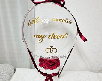 May include: A clear balloon shaped like a hot air balloon with the text "Will you complete my deen?" and a gold ring symbol. The balloon is tied with a burgundy ribbon and attached to a white box with a burgundy ribbon and a bouquet of red roses.