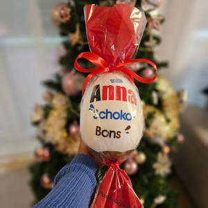Peut inclure: Un &oelig;uf en chocolat blanc envelopp&eacute; de cellophane rouge avec un ruban rouge. L'&oelig;uf est d&eacute;cor&eacute; des mots "Anna", "Schoko" et "Bons".