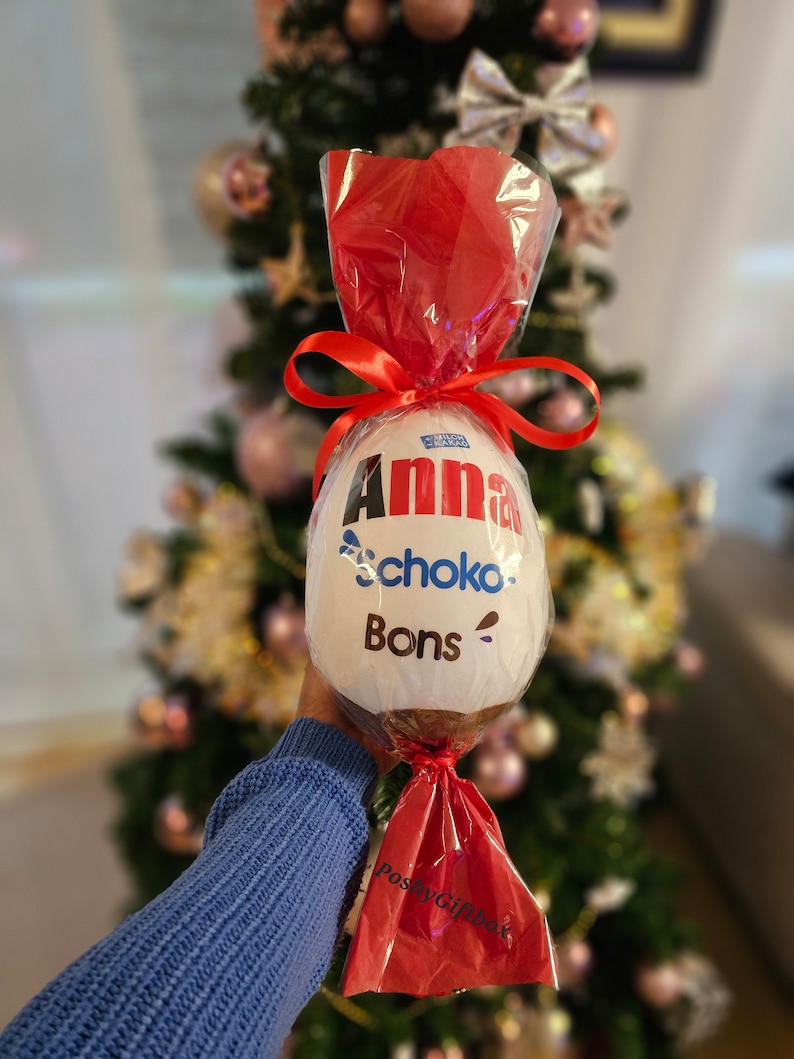Peut inclure: Un &oelig;uf en chocolat blanc envelopp&eacute; de cellophane rouge avec un ruban rouge. L'&oelig;uf est d&eacute;cor&eacute; des mots "Anna", "Schoko" et "Bons".