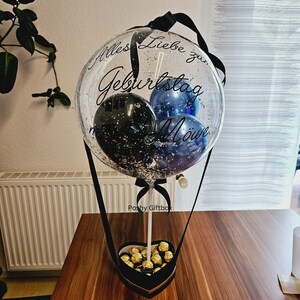 Op de afbeelding: Een doorzichtige ballon gevuld met zilveren confetti en kleinere blauwe en zwarte ballonnen, voorzien van de zwarte tekst "Alles Liebe zum Geburtstag M&ouml;we". De ballon is bevestigd aan een hartvormige zwarte doos gevuld met goudkleurige chocolaatjes.