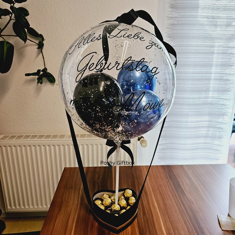 Op de afbeelding: Een doorzichtige ballon gevuld met zilveren confetti en kleinere blauwe en zwarte ballonnen, voorzien van de zwarte tekst "Alles Liebe zum Geburtstag M&ouml;we". De ballon is bevestigd aan een hartvormige zwarte doos gevuld met goudkleurige chocolaatjes.