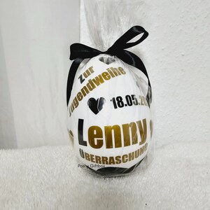May include: White egg-shaped gift wrapped in clear cellophane and tied with a black ribbon. The egg features gold and black text that reads "Zur Jugendweihe", "Lenny", "&Uuml;berraschung", a black heart, and the date "18.05.20".