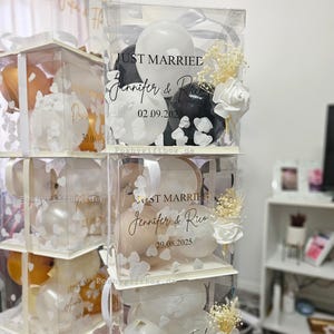 May include: Clear acrylic boxes stacked, each filled with balloons and floral accents. The boxes are decorated with the words "Just Married" and names, along with dates. White ribbons and bows adorn the boxes, creating a festive display.