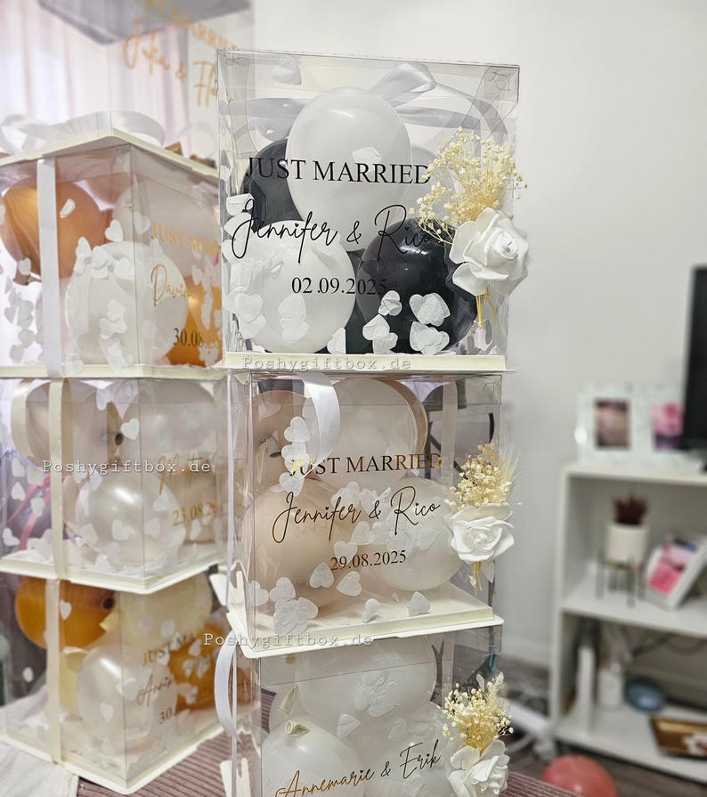 May include: Clear acrylic boxes stacked, each filled with balloons and floral accents. The boxes are decorated with the words "Just Married" and names, along with dates. White ribbons and bows adorn the boxes, creating a festive display.