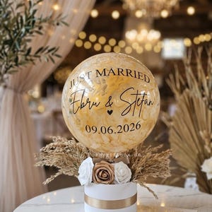 以下が含まれることがあります： 金色の花びらが入った透明なバルーン。黒い文字で「JUST MARRIED」「Ebru & Stefan」「09.06.2026」と書かれています。バルーンは、白と金のバラ、ドライパンパスグラスで飾られた白い円筒形の箱の上に設置されています。